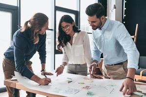 Another project. Group of young confident business people working together and smiling while man writing on blueprint in the office photo