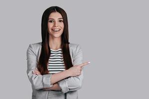 Pointing copy space. Beautiful young woman in smart casual wear keeping arms crossed and looking at camera with smile while standing against grey background photo