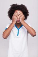I see nothing. Teenage African boy covering eyes by hands while standing isolated on grey background photo