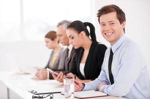 Business seminar. Side view of confident business people sitting in a row at the table while one man looking at camera and smiling photo