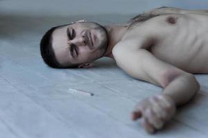 Feeling so badly. Young shirtless man lying on the floor and keeping eyes closed while syringe with narcotic laying near him photo