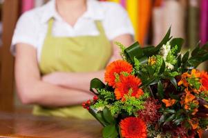 en la floristería. imagen recortada de una joven rubia en delantal con los brazos cruzados mientras un ramo de flores se pone en primer plano foto