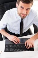 Confident manager. Top view of handsome young man in formalwear working on laptop and smiling while sitting at his working place photo