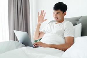 Asian man in white t-shirt laying on bed using laptop for video call waving hand making hello gesture. photo