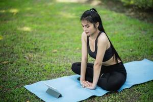 Asian sport fitness woman learning by doing a yoga according to a video clip or  video conferrence with teacher. Asian woman using tablet and doing yoga in park. Yoga study learning online concept. photo