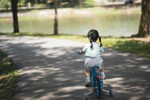 vista lateral trasera de una niña asiática aprendiendo a andar en bicicleta en el jardín del parque. concepto de educación para niños que practican ciclismo en el parque, concepto de deporte para bebés. foto