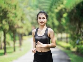 una deportista asiática se ajusta al cuerpo con ropa deportiva negra y sonríe mientras corre o trota en el jardín, tiene más árboles y aire limpio, hace ejercicio en el parque. concepto de carrera deportiva saludable. foto