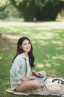 Asian woman sitting on picnic blanket and lawn at park working on laptop. Asian female using laptop while sitting under a tree at park with bright sunlight. Work from anywhere concept. photo