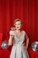 mature stylish elegant woman in cocktail dress with glass of sparkling wine with presents on red background. Party, fashion, celebration, anti age concept photo