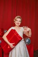 mujer madura y elegante con vestido de cóctel con una copa de vino espumoso con una caja de regalo en las manos. fiesta, moda, celebración, concepto anti edad. foto