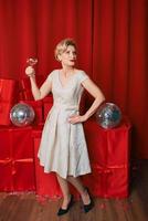 mature stylish elegant woman in cocktail dress with glass of sparkling wine with presents on red background. Party, fashion, celebration, anti age concept photo