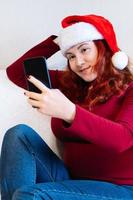 Young red-haired woman in Santa hat sits on couch at home and uses mobile phone to videochat. Online greeting with New Year and Christmas. photo