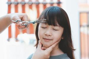 manos de mujer para cortar el pelo delantero o flequillo para una linda chica asiática en casa. las madres están felices de cortar el pelo de sus hijos. concepto de cuidado del cabello foto