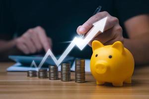 Businessman holding a coin in a piggy bank with graphs and arrows showing charts on his desk. concept of money growth and business finance, successful investment strategy photo