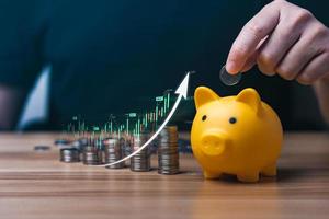 Businessman holding a coin in a piggy bank with graphs and arrows showing charts on his desk. concept of money growth and business finance, successful investment strategy photo