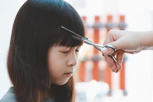 la mano femenina corta el cabello para una chica asiática con tijeras y peine. concepto de cuidado del cabello foto
