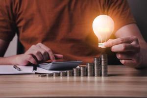 businessman hand holding lightbulb icon with coins stacking and calculator on desk. collecting money, financial planning, save money for the future, saving energy photo