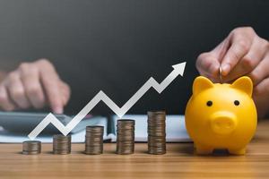Businessman holding a coin in a piggy bank with graphs and arrows showing charts on his desk. concept of money growth and business finance, successful investment strategy photo