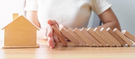 la mano de la mujer deja de caer bloques de madera y protege el modelo de la casa. crisis, negocio de caída, riesgo, recesión económica, desarrollador, bienes raíces y concepto de propiedad foto