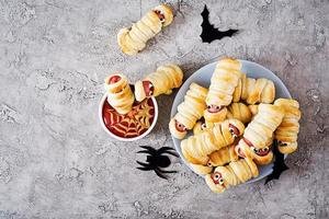 Scary sausage mummies in dough with funny eyes on table. Funny decoration. Halloween food. Top view. Flat lay photo