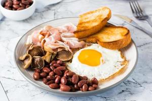 English breakfast with fried egg, bacon, beans, mushrooms and toast on a plate on the table.  Close-up photo