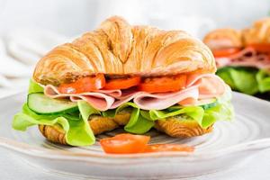 Fresh croissant sandwich with ham, vegetables and lettuce on a plate. Close-up. photo