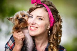 beautiful young girl in a plaid shirt and short denim shorts in pin-up style in forest with yorkshire terrier small dog in her arms