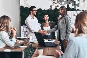 Glad to work with you Young modern men in smart casual wear shaking hands and smiling while standing in the creative office photo