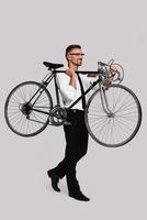 tome una bicicleta a lo largo de un joven con traje completo que lleva su bicicleta mientras camina contra un fondo gris foto