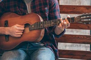 Playing perfect chord. Close-up part of young man playing guitar while sitting at windowsill photo