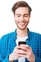 My gadget is my helper. Happy young man in shirt holding mobile phone and smiling while standing against white background photo