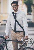 Getting to work. Cheerful young man in glasses looking at camera with smile and holding hand on his bicycle while standing outdoors photo