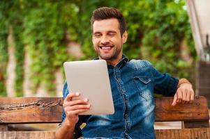 Relaxing with his new tablet. Happy young man holding digital tablet and smiling while sitting on the wooden bench outdoors photo