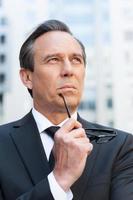 Thinking about business solutions. Thoughtful mature man in formalwear holding glasses near face and looking away while standing outdoors photo