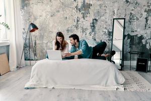 Looking for a movie to watch. Beautiful young couple using computer while spending time in bed at home photo
