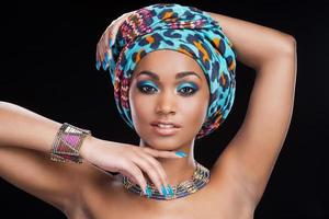 In traditional African style. Beautiful African woman in headscarf and jewelry posing against black background and looking at camera photo