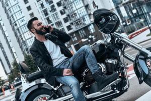 Good to hear you Handsome young man in leather jacket talking on the phone and smiling while sitting on the motorbike outdoors photo