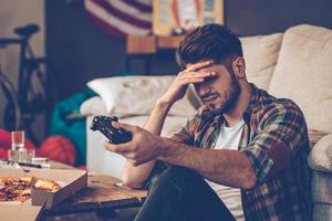 Game over. Frustrated young man holding joystick and keeping his hand on forehead while sitting on the floor in messy room after party photo