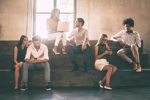 Students life. Group of cheerful young people communicting while holding different gadgets and sitting close to each other on steps photo