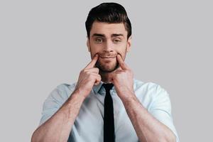 Nothing cheers him up. Sad young man in white shirt and tie trying to smile by touching his face with fingers and looking at camera while standing against grey background photo