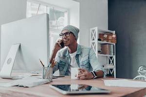 Good talk with client. Handsome young man in eyewear talking on smart phone and smiling while sitting at his working place in creative office photo