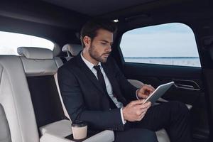 teniendo en cuenta el siguiente paso. un joven apuesto con traje completo que trabaja con una tableta digital mientras está sentado en el auto foto