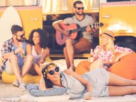 Summer time with friends. Cheerful young woman smiling at camera while lying near retro minivan with friends in the background photo