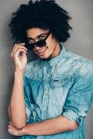 Cheerful and stylish.Cheerful young African man adjusting his sunglasses and looking at camerawith smile while standing against grey background photo