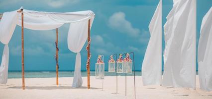 decoración romántica de bodas en la playa de ensueño, arena blanca y cielo azul nublado. pareja ama la luna de miel y la celebración del aniversario, un ambiente soleado de color pastel suave, flores y velas bajo cortinas blancas foto