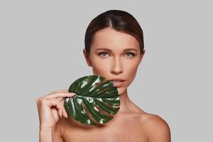 She is perfection. Attractive young woman looking at camera and covering with leaf while standing against grey background photo