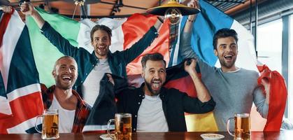 Excited young men covered in international flags drinking beer and watching sport game while sitting in the pub photo