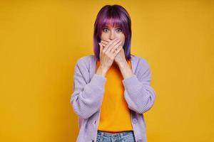 Shocked young woman covering mouth with hands while standing against yellow background photo