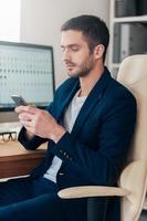Typing business message. Side view of confident young man holding smart phone and looking at it while sitting at his working place in office photo