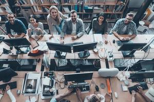 Young and successful. Top view of group of young business people in smart casual wear working together and smiling while sitting at the large office desk photo